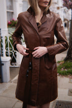 Woman wearing a brown leather coat standing on a street with buildings and trees in the background.